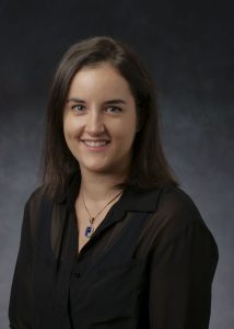 A professional headshot of Dr. Eryn Cangi, a white woman with shoulder-length brown hair. She is wearing a sheer black button down over a black tank top and a necklace.