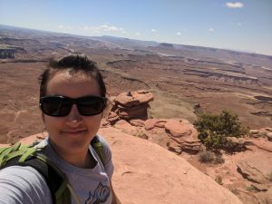 A woman wearing sunglasses with brown hair pulled into a ponytail is taking a selfie in front of a staggeringly large desert canyon. The scene comprises red rock in the foreground, with a juniper tree in the midground down a slope, and blue sky in the distance. 