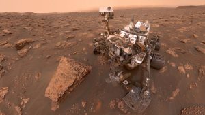 the curiosity rover selfie from Mars. Curiosity is viewed on a red, dusty desert strewn with rocks, with a bright salmon sky beyond.